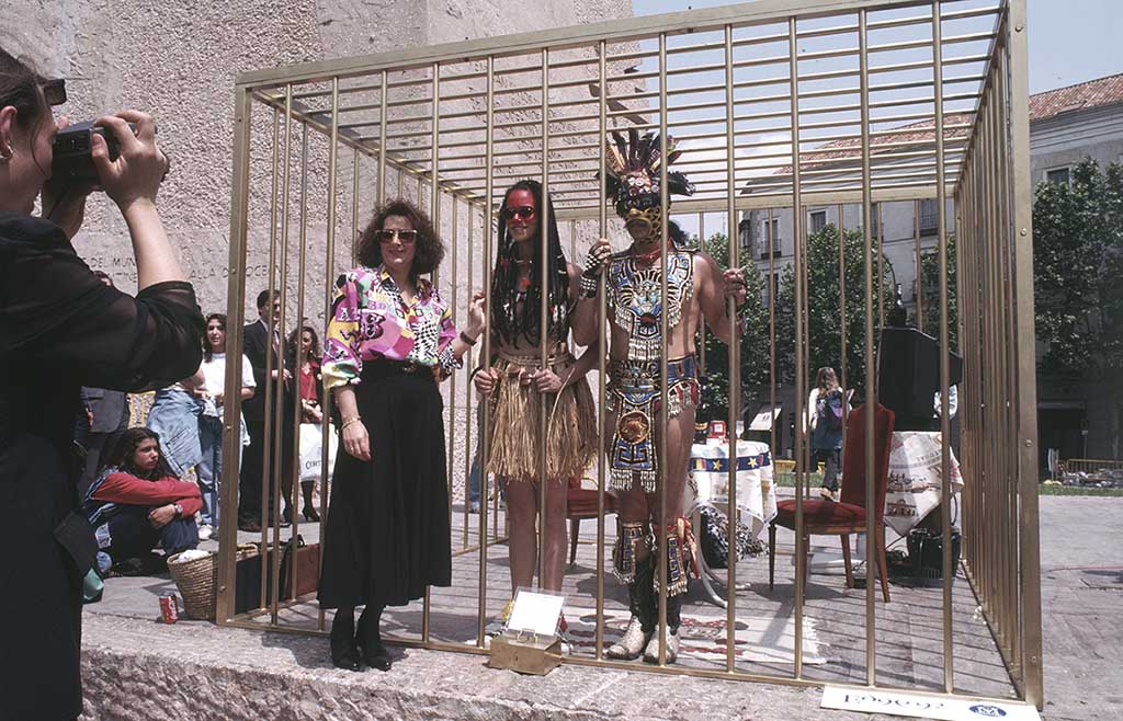 Two people dressed as Amerindians standing in a large gold colored cage. A woman poses in front of the cage while another woman photographs them.